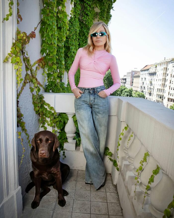 Donatella Versace in a pink top and jeans on a balcony with greenery, showcasing her stunning transformation.