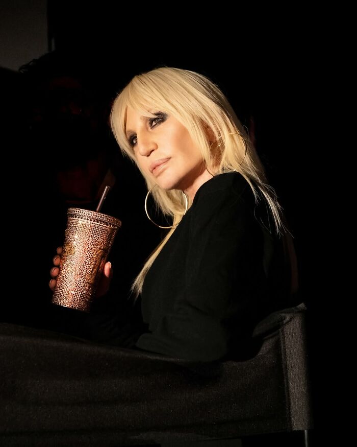 Donatella Versace with blonde hair and hoop earrings holding a sparkling cup, showcasing her stunning transformation.