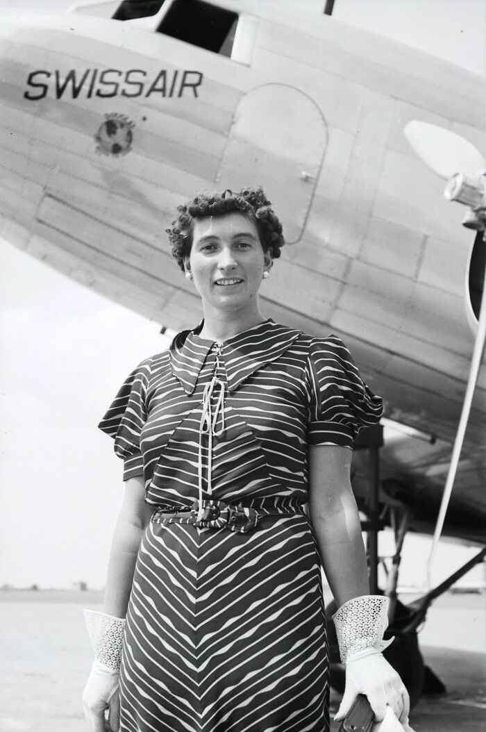 Vintage black and white photo of a glamorous female flight attendant standing in front of a Swissair airplane.