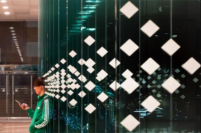 Young man in green jacket leaning against glass wall with white geometric shapes, showing street coincidences caught on camera.