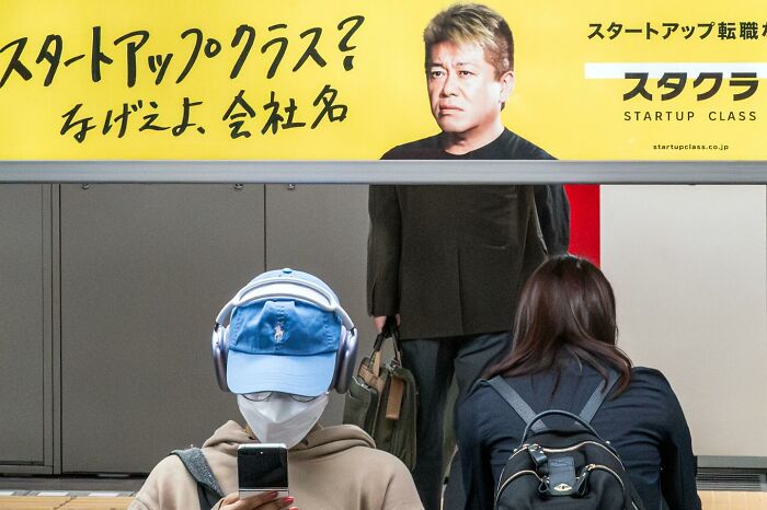 Person wearing headphones and a mask looking at their phone under a yellow billboard in a street coincidence scene.