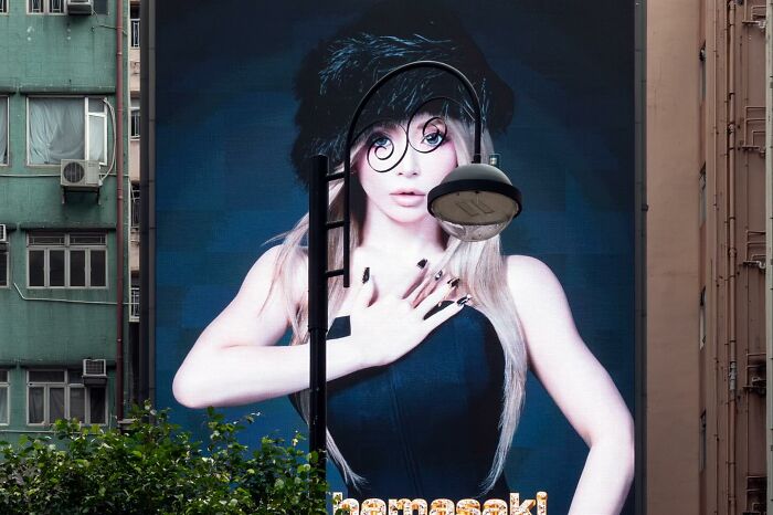 Street coincidences captured with a lamp post perfectly aligned over a woman's face on a large urban billboard.