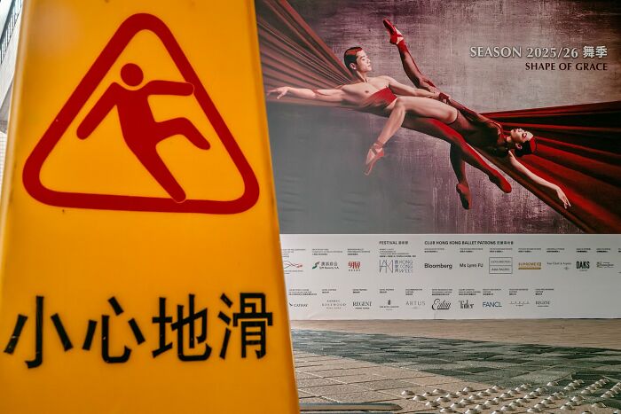 Yellow caution sign showing slipping figure in front of a ballet advertisement, illustrating street coincidences caught on camera.