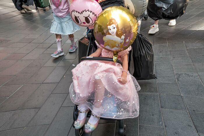 Child in a pink dress holding a princess balloon, blending with balloons in a street coincidence captured on camera.