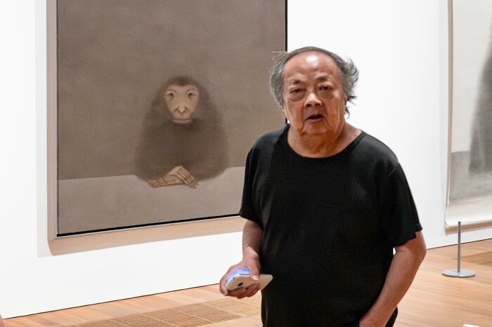 Man in black shirt standing in front of art that creates a street coincidence with a primate figure in the background.
