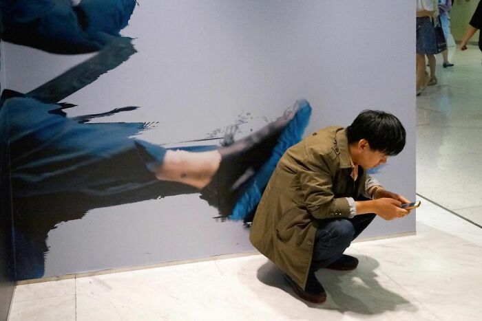 Man crouching and looking at phone aligned with street art showing large kicking feet, street coincidences captured on camera.