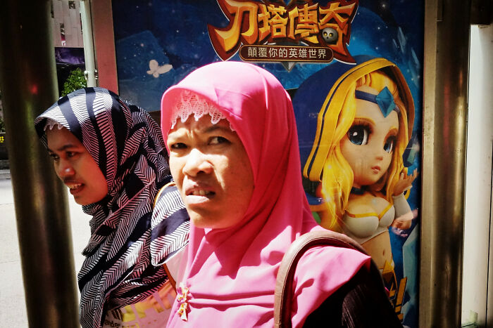 Two women wearing hijabs stand near a colorful animated character poster in a vibrant street coincidence scene.