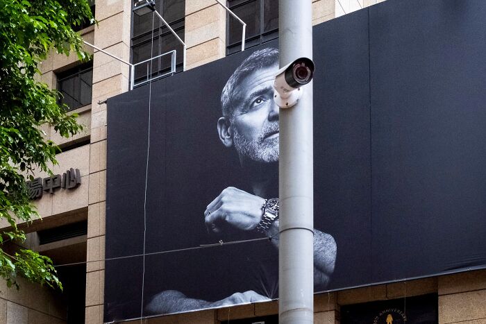 Black and white street coincidence image with a security camera aligned perfectly with a man’s eye on a billboard.
