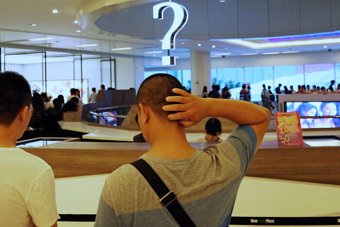 Man scratching head underneath illuminated question mark sign in a crowded indoor space, illustrating street coincidences.