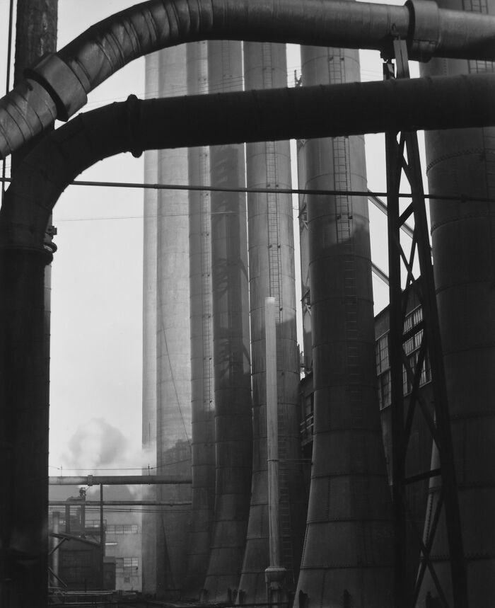 Black and white image of industrial smokestacks and large pipes highlighting 20th century industrial development.