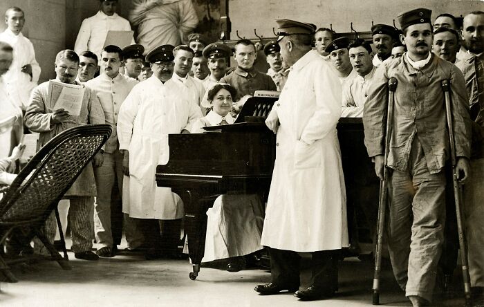 WWI soldiers and medical staff gathered around a piano, showing daily life beyond the WWI trenches in a hospital setting.