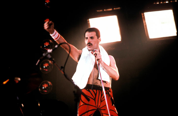 Freddie Mercury performing on stage with microphone, spotlight highlighting singer during energetic live concert.