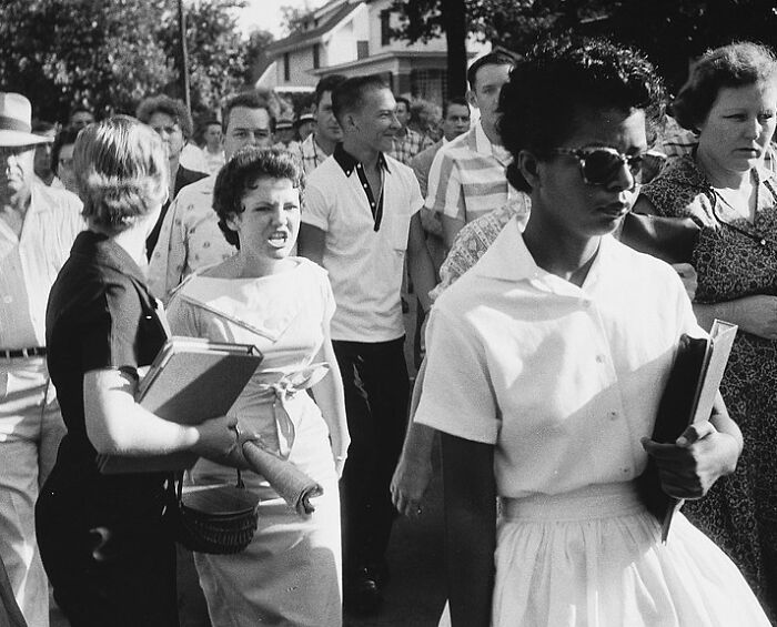 Black and white photo showing a tense crowd walking with a determined African American woman holding books in the 20th century.