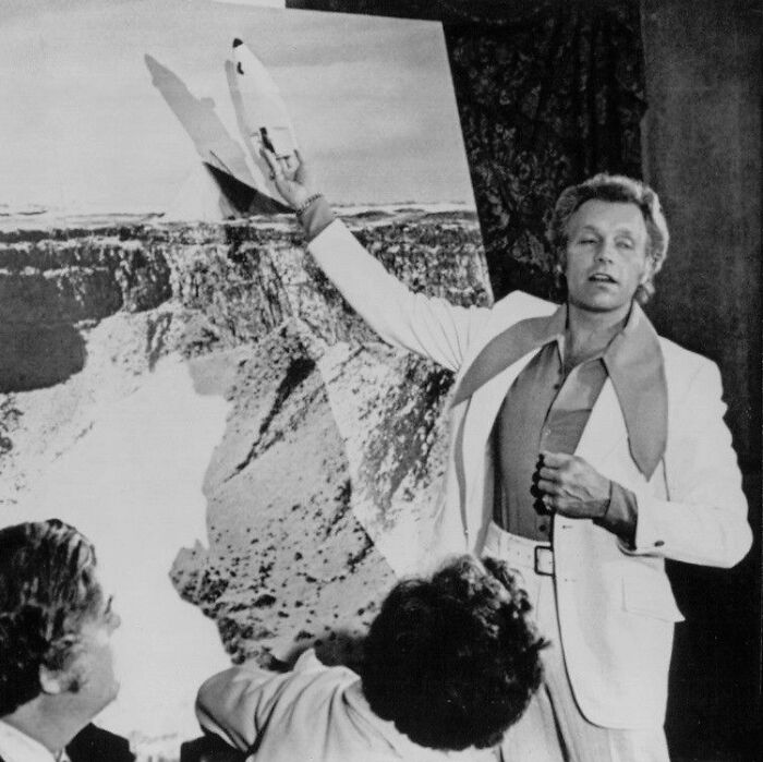 Man in white suit pointing to a space shuttle image while sharing new facts for those who like learning new things.