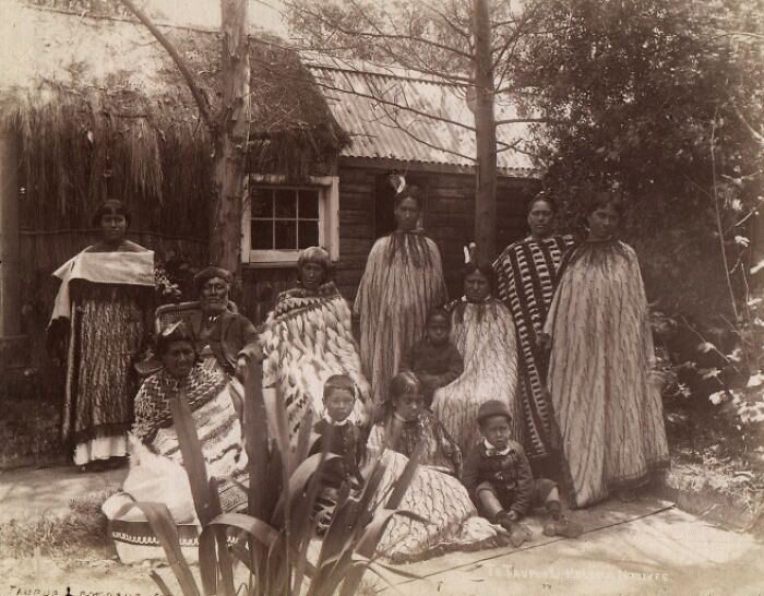 Group portrait of Māori people wearing traditional cloaks outside a historic rural dwelling, reflecting Māori life a century ago.