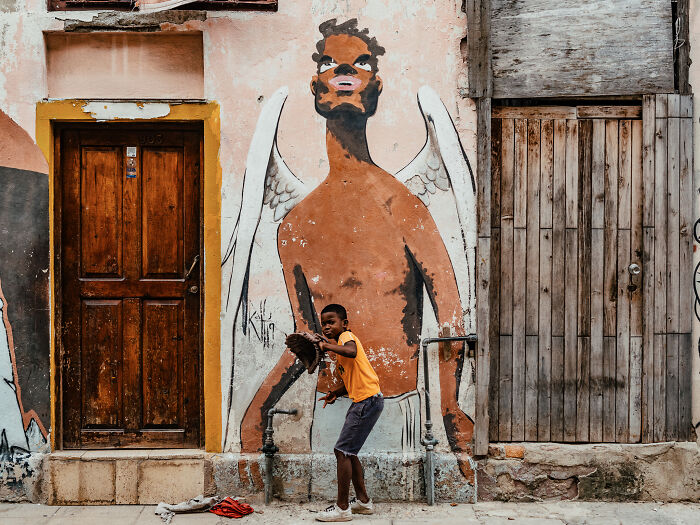 Boy playing baseball in front of a mural on a weathered street wall, capturing street photography life moments.