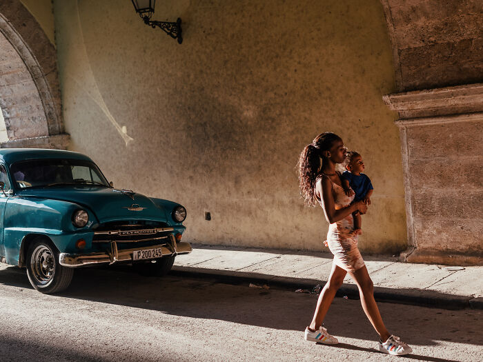 Woman walking while carrying a child past a vintage car under an archway in a street photography shot capturing life.