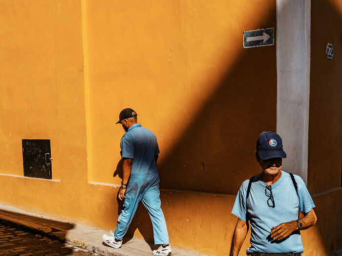 Two men in blue clothing walking near a sunlit yellow wall, capturing street photography shots that depict life realistically.