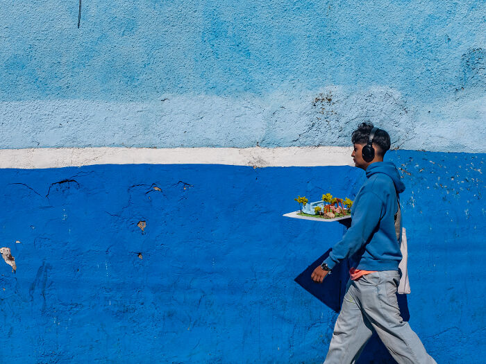 Young man walking with headphones and plants against a textured blue wall in street photography shots capturing life.