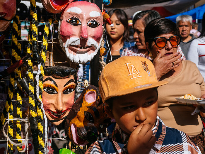 Child wearing yellow LA cap among colorful street masks with crowd in background, vibrant street photography capturing life.
