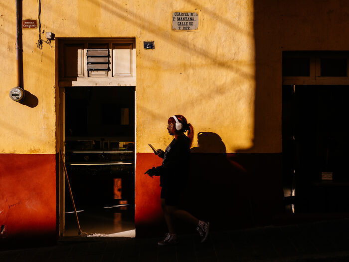 Person walking past vibrant yellow and red wall, wearing headphones and holding a phone in a street photography shot.