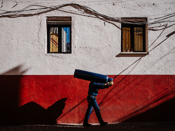 Man carrying a large cylinder walking past a red and white wall in a vibrant street photography shot capturing life.