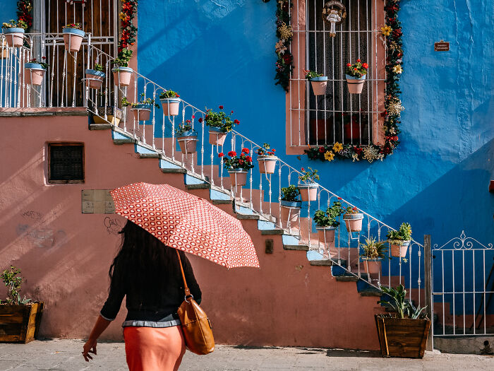 Woman holding patterned umbrella walking past colorful building with stairs and potted plants in vibrant street photography shot.