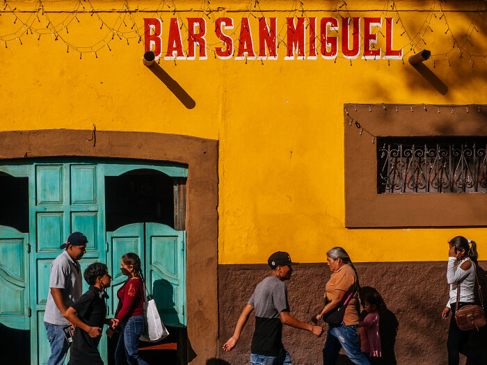 Colorful street photography shot of pedestrians walking past a vibrant yellow wall with Bar San Miguel sign outdoors.