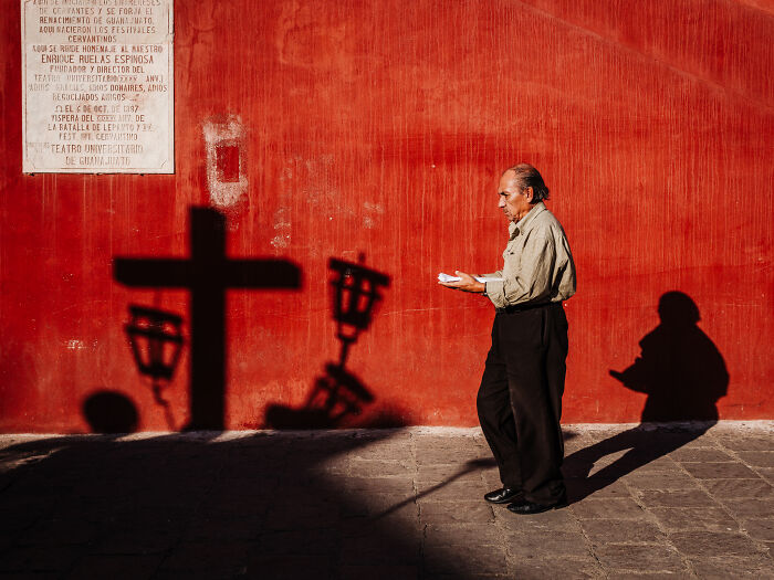 Man walking past red wall with cross shadows in a street photography shot capturing life as it really is by Federico Borobio