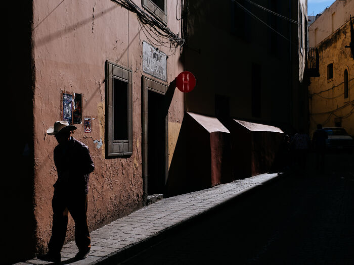 Man in a hat walking along a sunlit street corner, captured in a striking street photography shot showing urban life.