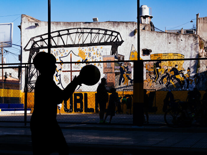 Silhouette of a child playing with a ball in front of a colorful mural in vibrant street photography capturing life realistically.