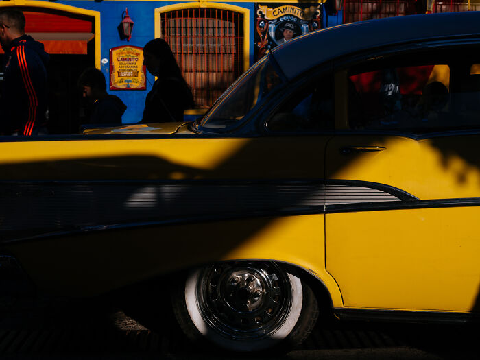 Yellow vintage car with shadows and silhouettes of people beside a vibrant blue building in street photography.