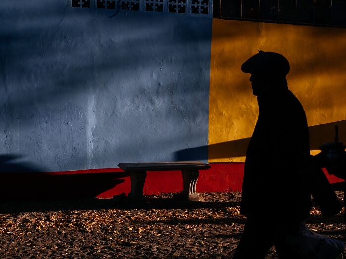 Silhouette of a person walking past a colorful wall with strong shadows in a street photography shot capturing life.