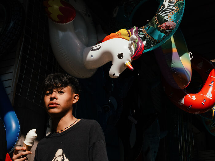 Young person holding ice cream cone under colorful inflatable unicorn, vibrant street photography shot capturing life realistically.