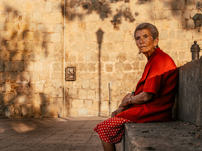 Elderly woman in red sweater and polka dot skirt sitting on a bench in warm light, street photography capturing real life.