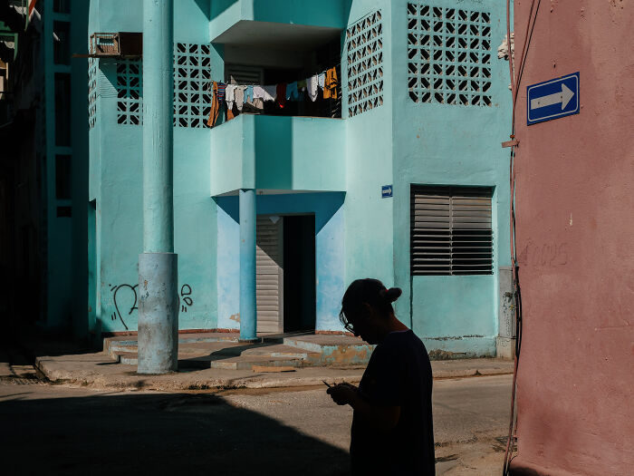 Person using phone in shadowed street corner with pastel blue building and laundry hanging, capturing authentic street photography life.