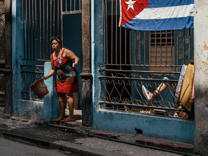 Woman in a red dress pouring water outside a blue building with a Cuban flag in a street photography shot.
