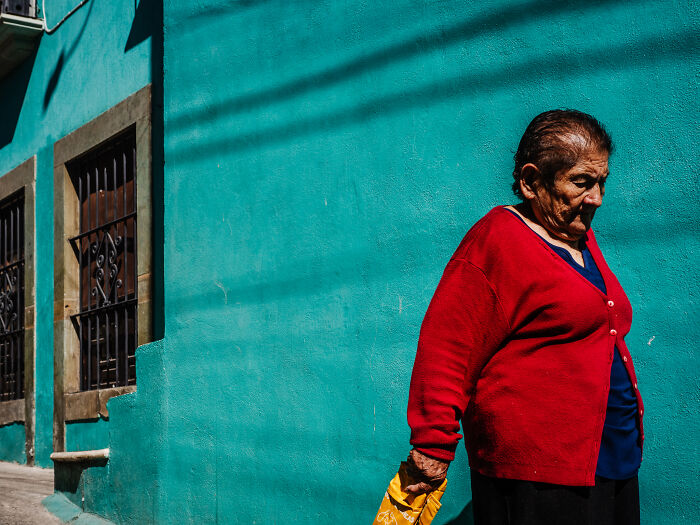 Elderly woman in a red sweater walking by a turquoise wall in a street photography shot capturing life realistically.