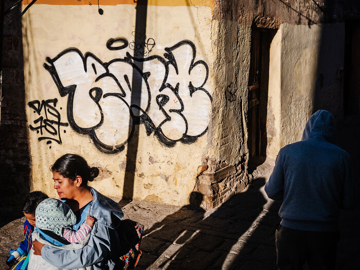 Street photography shot showing a woman carrying a child beside a graffiti wall with a person in a hoodie walking away.