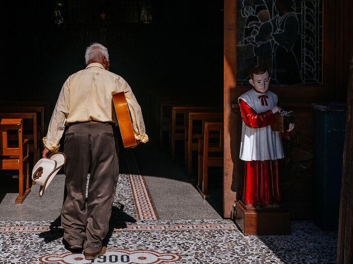 Elderly man holding a guitar and hat walking into a building, a statue stands near the entrance in a street photography shot.