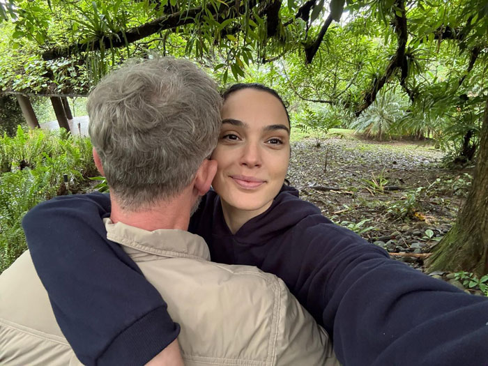 Gal Gadot smiling outdoors while hugging a man, surrounded by greenery and trees in a natural setting. Gal Gadot smiling outdoors while hugging a man, surrounded by greenery and trees in a natural setting.