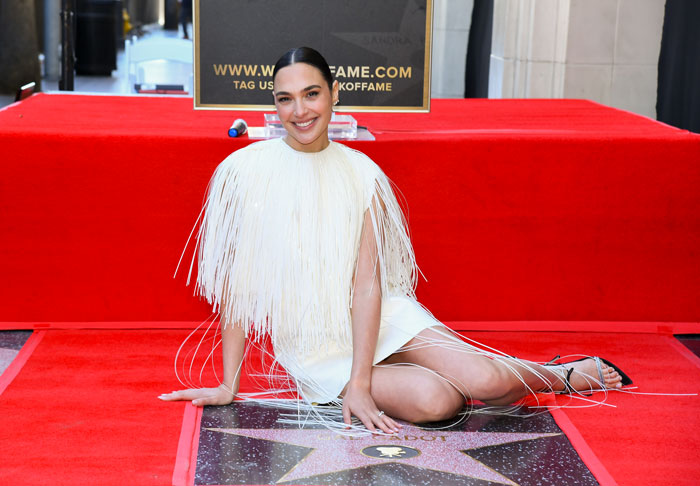 Gal Gadot sitting on Hollywood Walk of Fame star, smiling, wearing a white fringed dress and black sandals. Gal Gadot sitting on Hollywood Walk of Fame star, smiling, wearing a white fringed dress and black sandals.