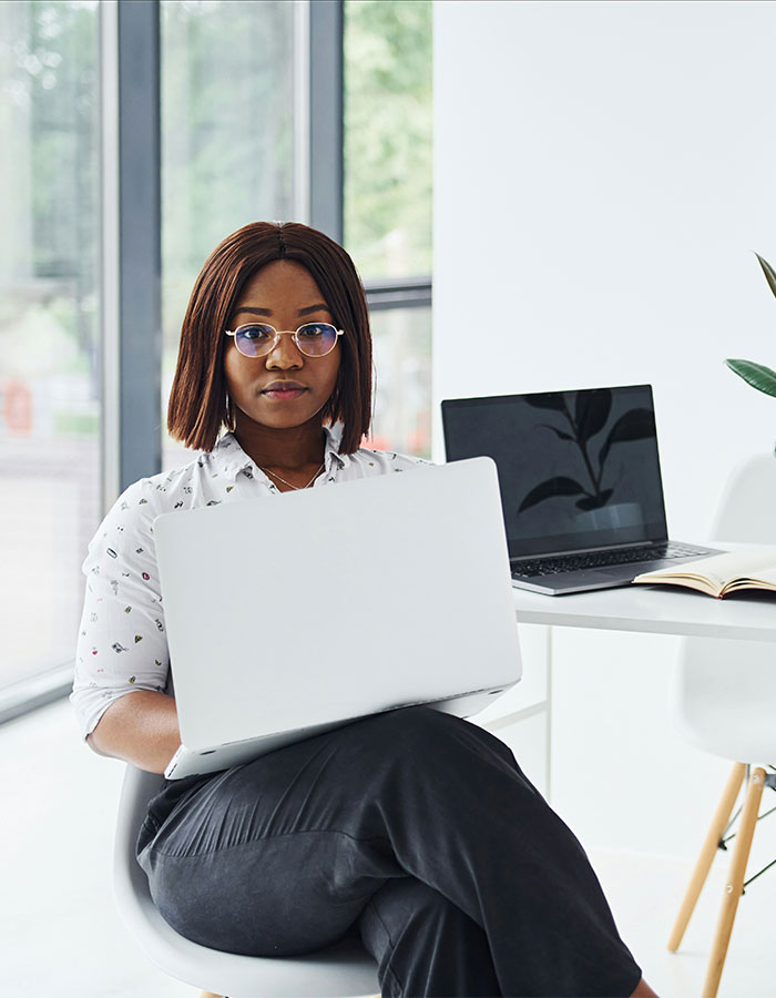Young Gen Z worker wearing glasses and office attire, sitting with a laptop in a modern workplace setting.