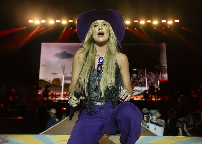Female singer in purple outfit and hat performing on stage, a memorable celebrity wardrobe malfunction moment.