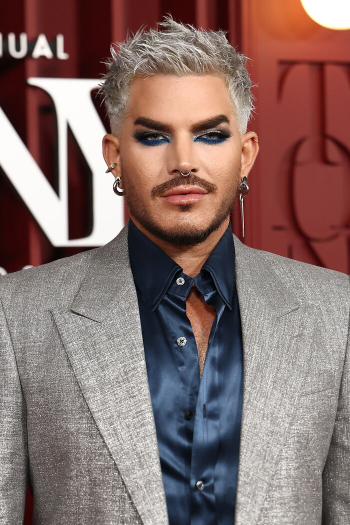 Adam Lambert with silver hair and bold makeup wearing a gray suit and blue shirt at a formal event. Adam Lambert with silver hair and bold makeup wearing a gray suit and blue shirt at a formal event.