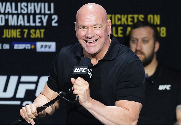 Dana White speaking at a UFC event, smiling and holding a microphone with UFC branding in the background.