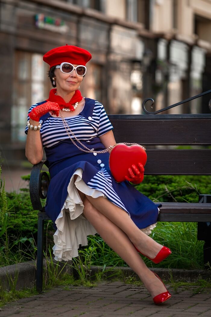 Woman in incredible cosplay outfit with red beret, gloves, and heart-shaped purse, sitting on a bench outdoors.