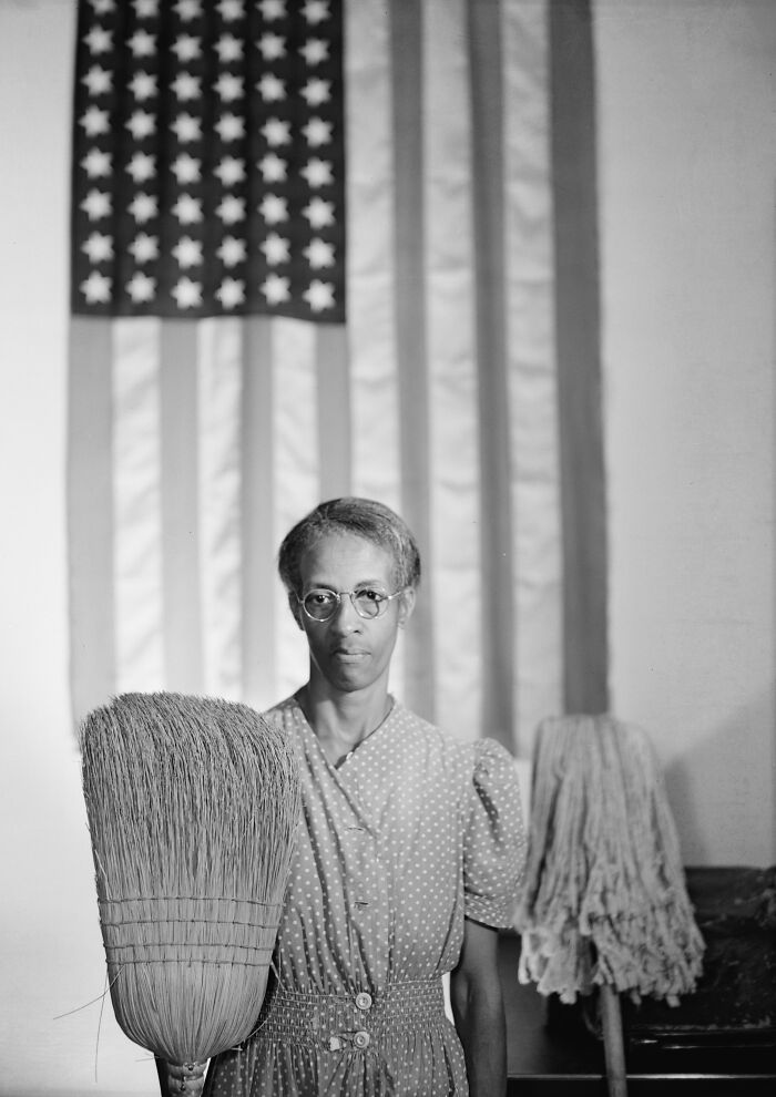 African American woman holding a broom standing in front of an American flag, symbolizing 20th century triumph and tragedy.