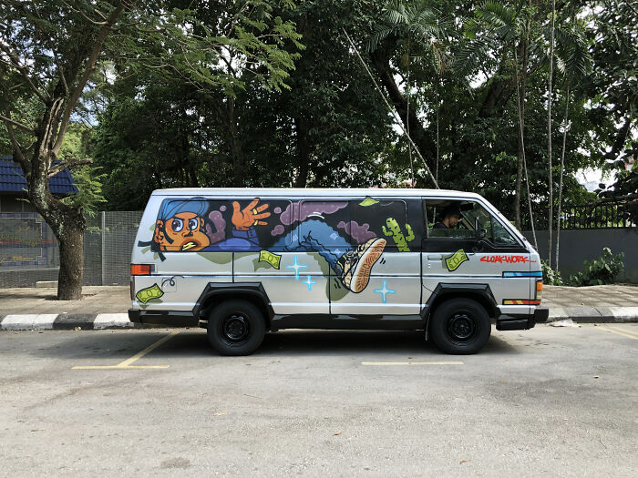 Van covered in colorful street art painting of a person and money, parked next to trees on a city street.