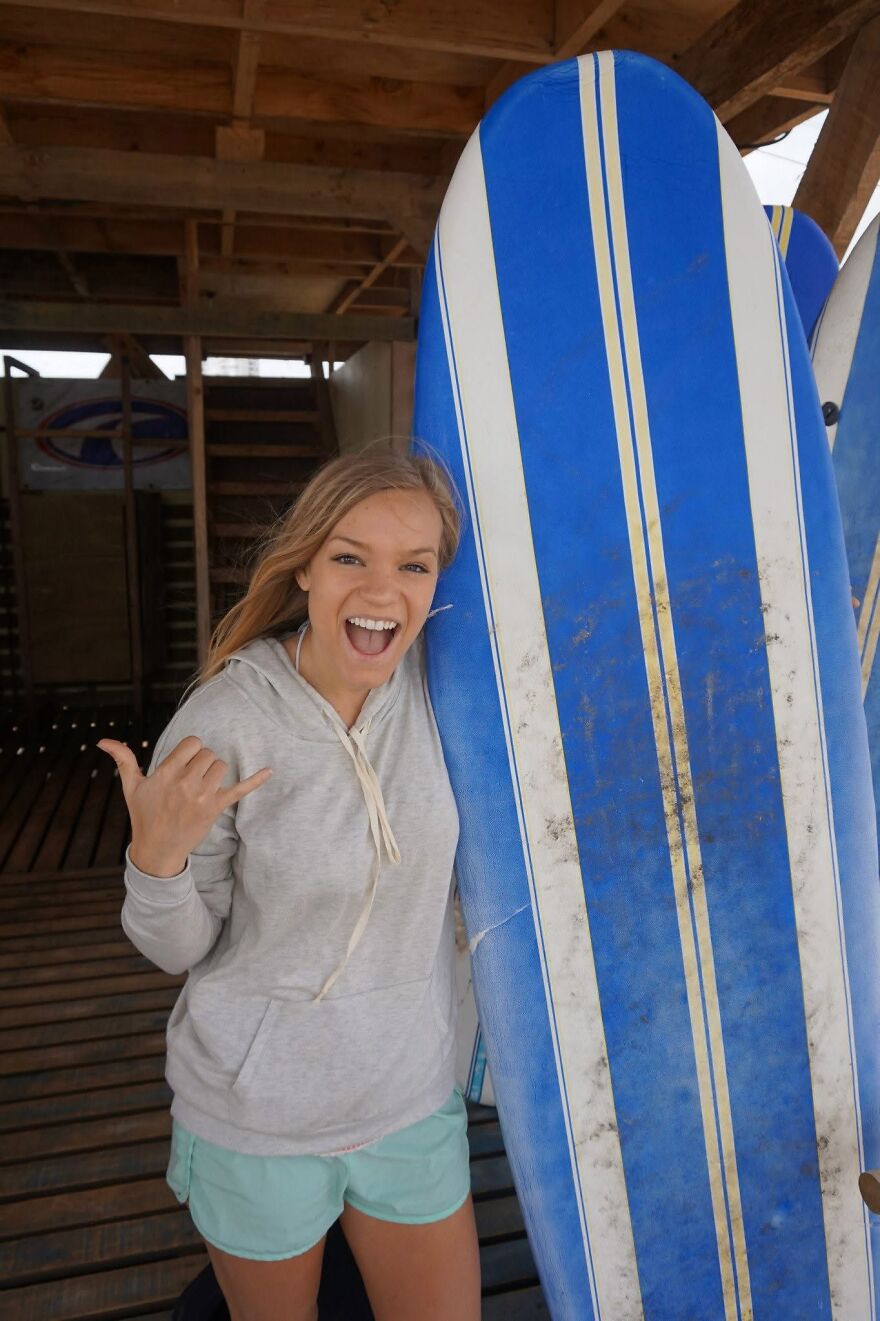 Young woman smiling and holding a blue surfboard, expressing excitement after a one-way flight to Australia adventure.